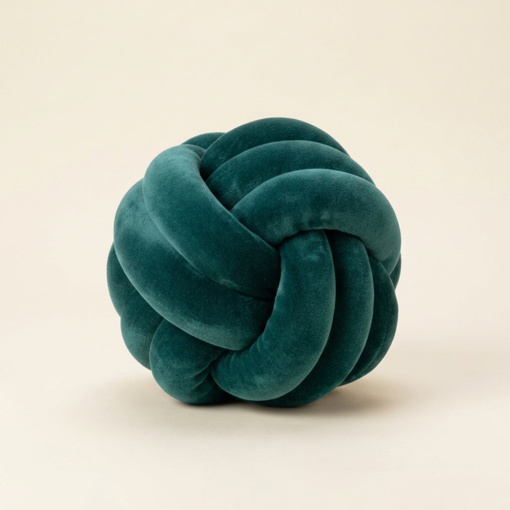 A dark teal velvet knot pillow centered on a warm off-white background.