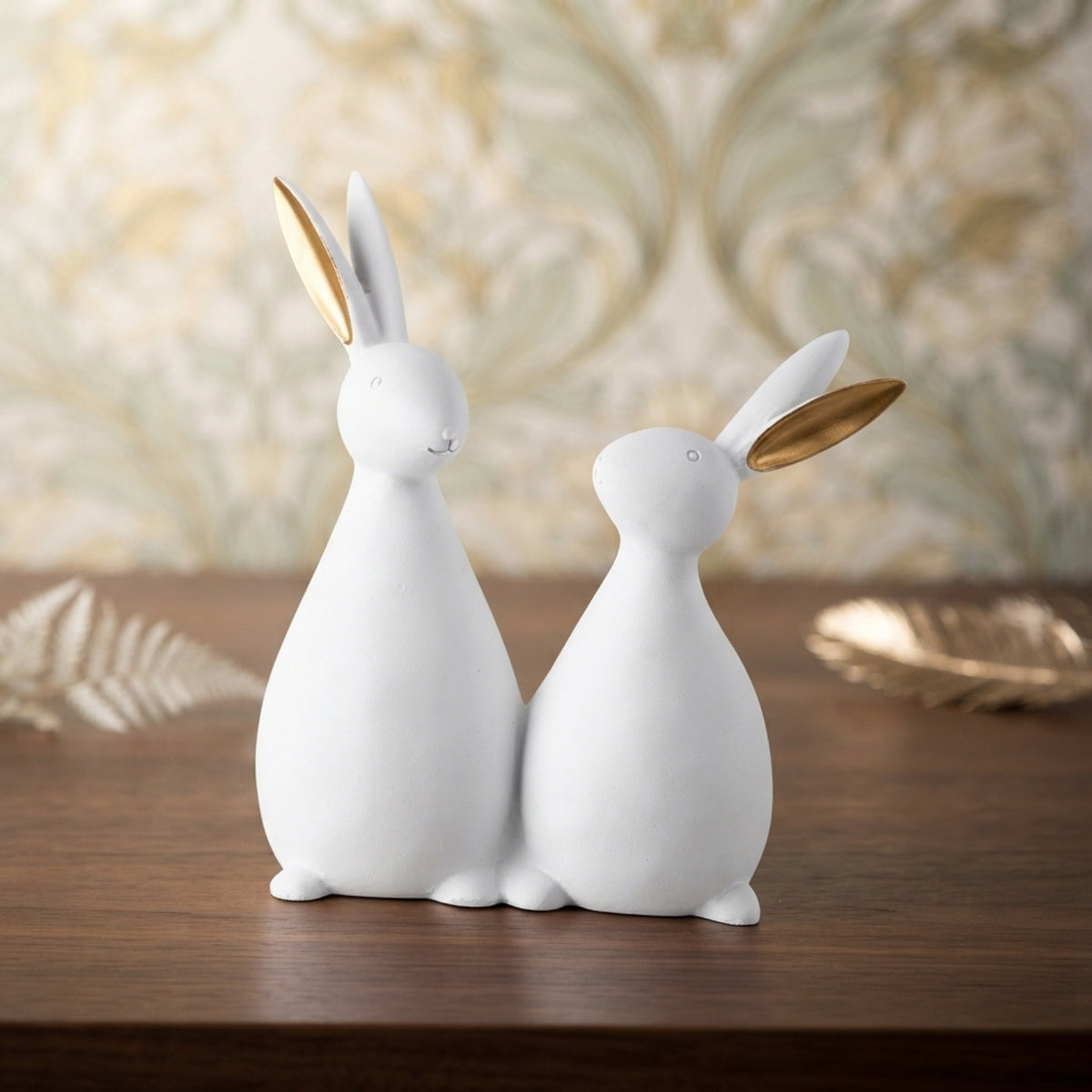 Two white rabbit figurines with metallic gold-leaf ears sitting on a dark wooden table, set against a softly blurred beige and gold patterned wallpaper background.