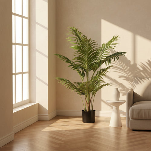 A tall artificial palm plant in a black pot, placed in a sunlit living room corner next to a white side table and a beige sofa.