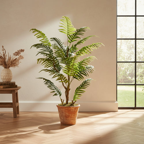 A potted artificial palm tree with lush green fronds in a terracotta-colored pot, placed on a light wood floor in a sunlit room next to a window.