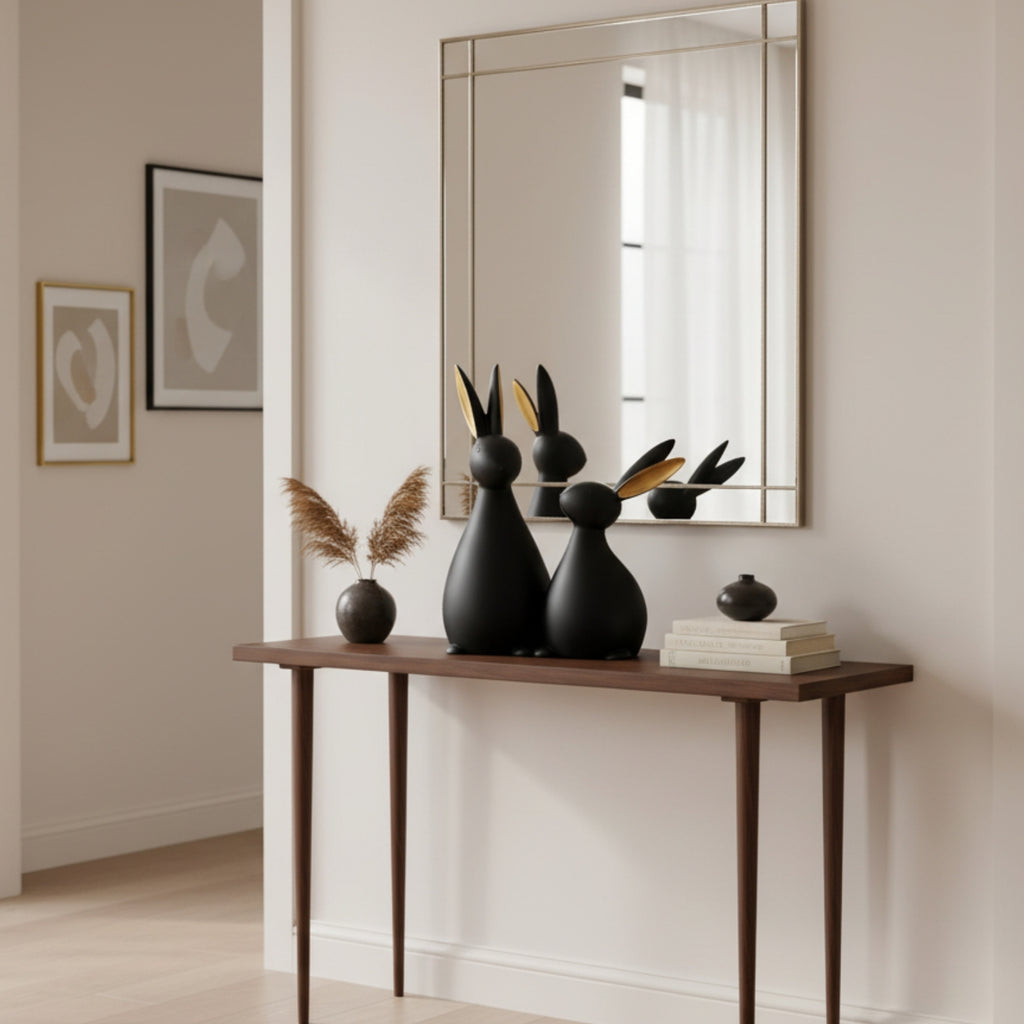 A styled interior scene showing a wooden console table against a cream wall, with a large rectangular mirror above it. On the table are two matte black rabbit-shaped decorative sculptures with gold leaf accents, a small vase with dried pampas grass, and stacked books.