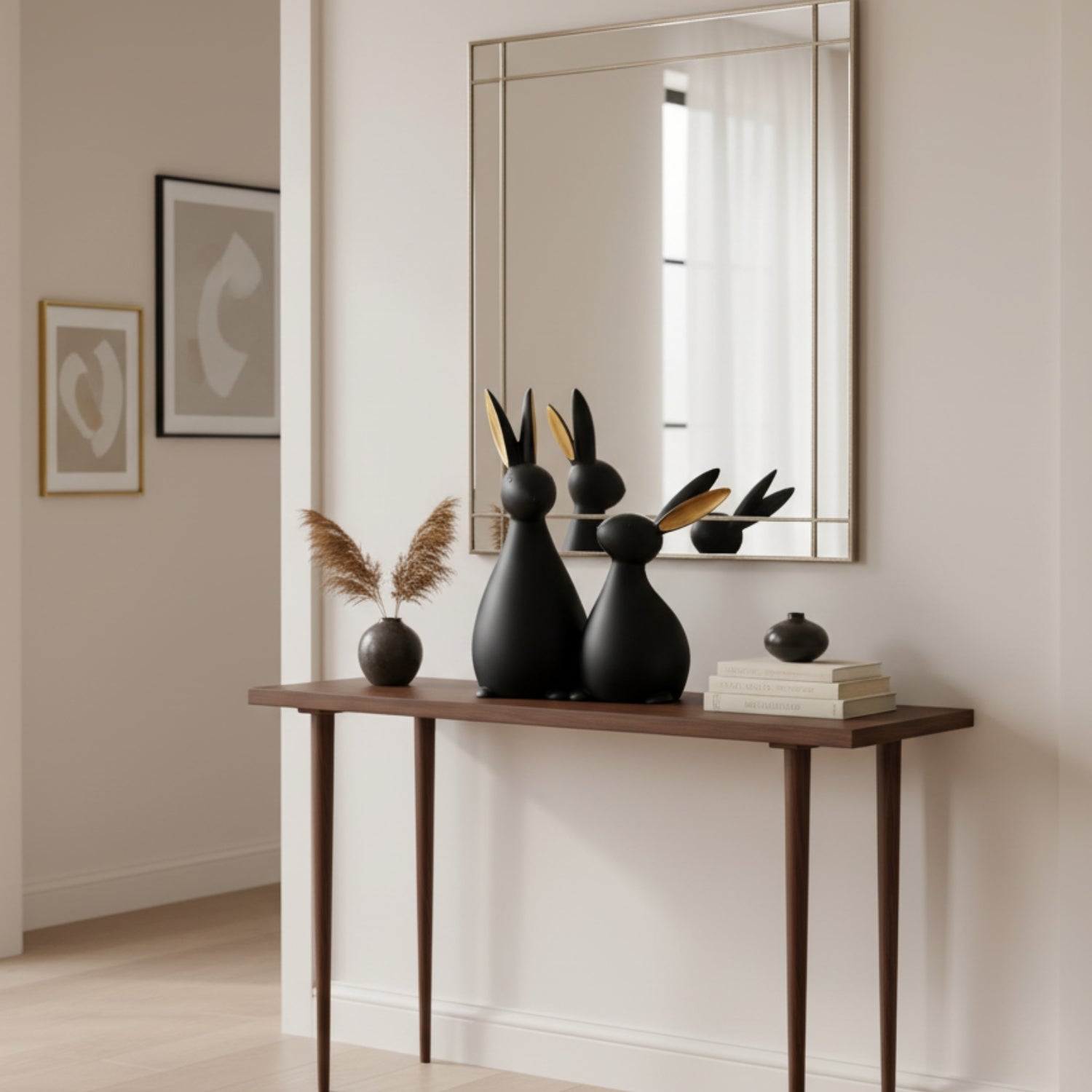 A styled interior scene showing a wooden console table against a cream wall, with a large rectangular mirror above it. On the table are two matte black rabbit-shaped decorative sculptures with gold leaf accents, a small vase with dried pampas grass, and stacked books.