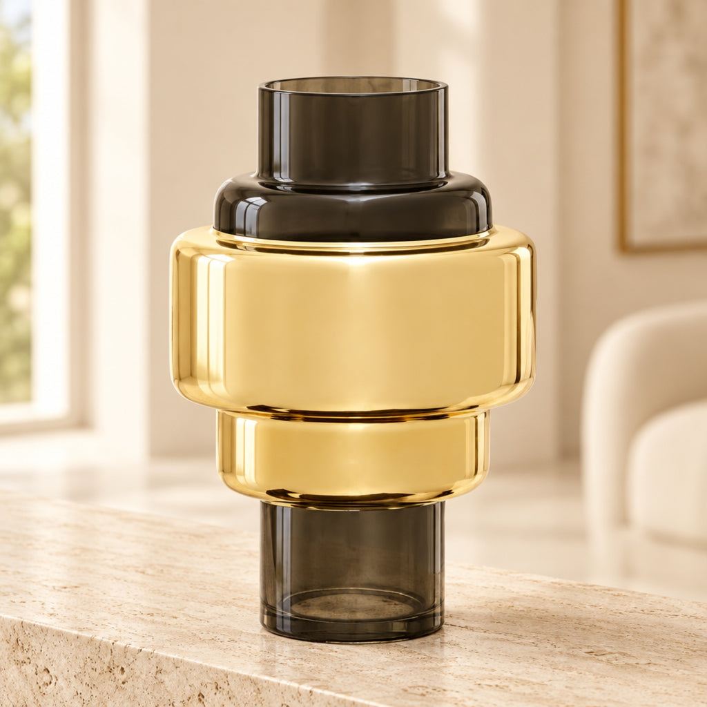 A modern stacked glass vase with a wide, reflective gold middle section and dark smoky gray top and bottom sections, displayed on a light stone surface in a softly blurred interior setting.