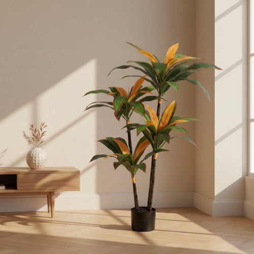 An artificial Dracaena massangeana houseplant with green and orange variegated leaves in a black pot, placed in a minimalist room with beige walls, light wood flooring, and a small wooden side table.
