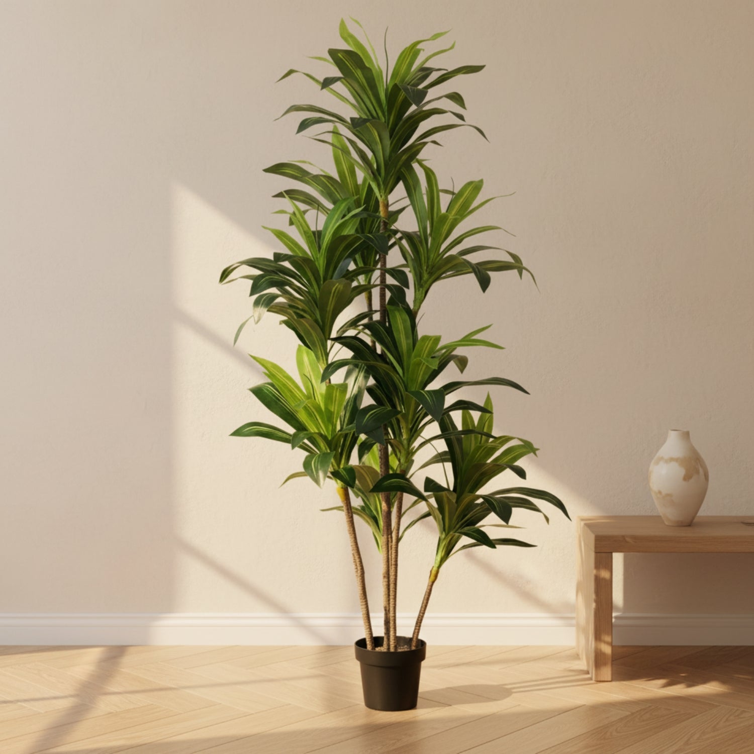 A tall artificial green plant in a black pot, styled in a minimalist room with a light wood floor, beige wall, and a small wooden side table with a ceramic vase.