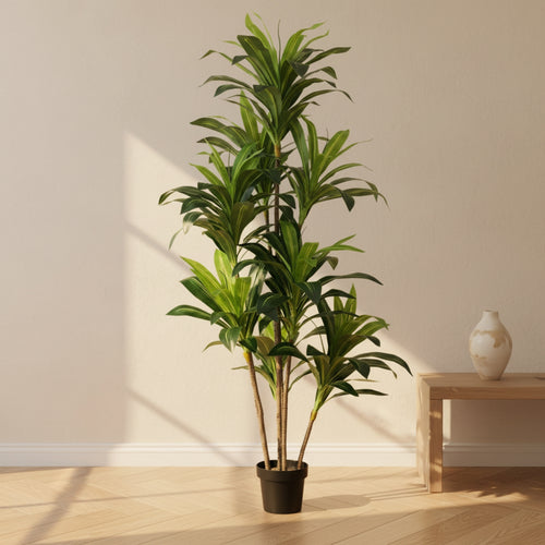 A tall artificial green plant in a black pot, styled in a minimalist room with a light wood floor, beige wall, and a small wooden side table with a ceramic vase.