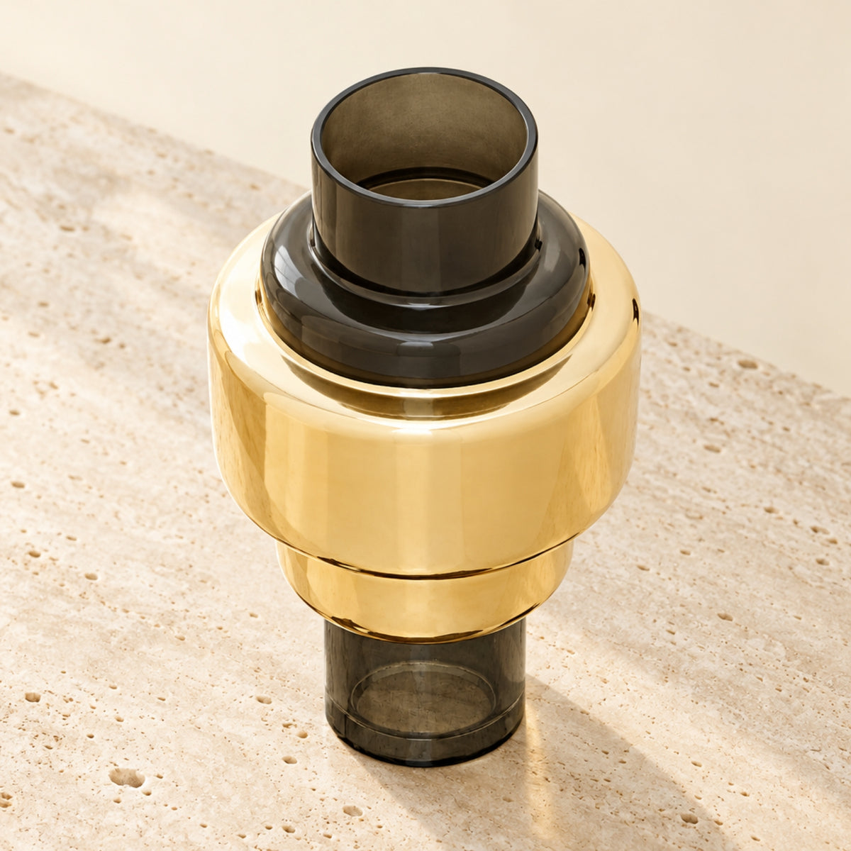 A modern cylindrical vase or diffuser with a polished gold-toned metallic body and dark smoky translucent glass top and base, photographed on a light stone surface against a warm neutral background.