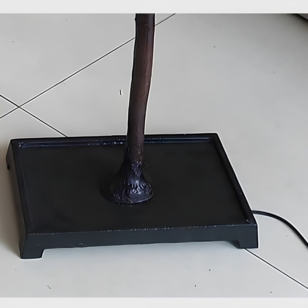 A close-up of a black rectangular floor lamp base with a dark brown central pole and a visible power cord on a light tiled floor.