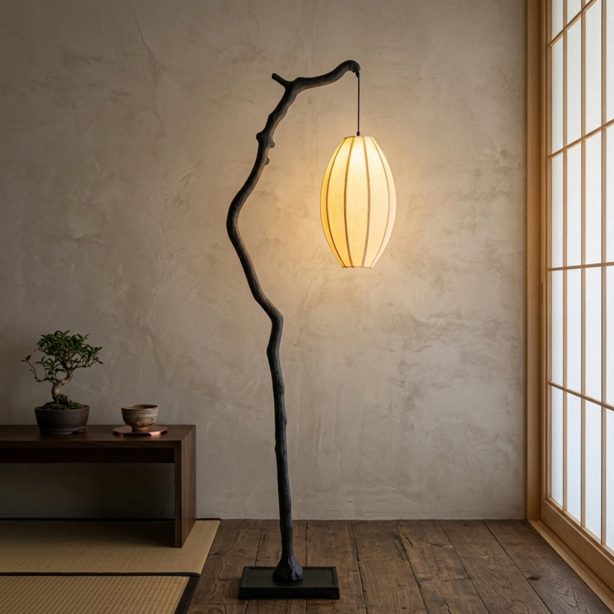 A modern floor lamp with a curved, branch-like black metal frame and a glowing warm white pleated paper shade, placed in a Japanese-style room with a textured beige wall, wooden floor, and shoji window.