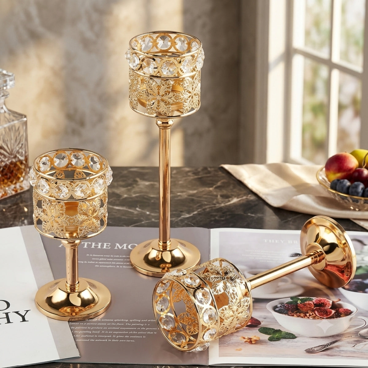 Three gold-toned candle holders with clear crystal rhinestone cups are arranged on a dark marble tabletop with open magazines and a fruit bowl in the background.