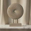 A decorative stone-like circular sculpture with a central hole, mounted on a rectangular base with two thin black legs. The top disc is light gray with thin copper-orange veining, set against a plain white background.