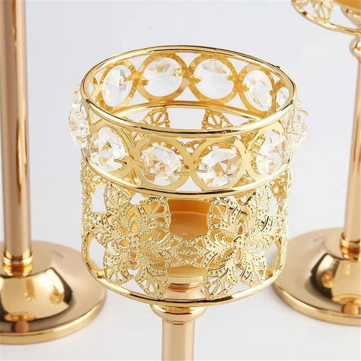 Gold metal candle holder with ornate filigree cutouts and clear faceted crystal accents, shown on a white background with part of another matching holder visible.