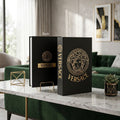 A black Versace hardcover book or notebook standing upright on a marble-topped table in a modern living room. The cover features a large gold circular Medusa emblem and the word VERSACE, with a smaller gold logo on the spine.