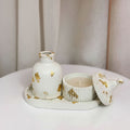 A white ceramic decorative set on a round tray, featuring a ribbed bottle-shaped vessel, a matching ribbed candle holder with lid, and scattered gold leaf accents, displayed on a white table against a soft beige curtain background.