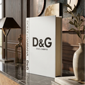 A white Dolce & Gabbana D&G book standing upright on a polished stone console table in a warm, wood-paneled interior. The book spine and cover are visible, with the bold black D&G logo and brand name printed on the front and spine.