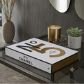 A Chanel No. 5 perfume box sits on a glass-top table in a modern interior setting with a gray concrete wall, decorative mirrors, a vase with dried grass, and a small succulent.